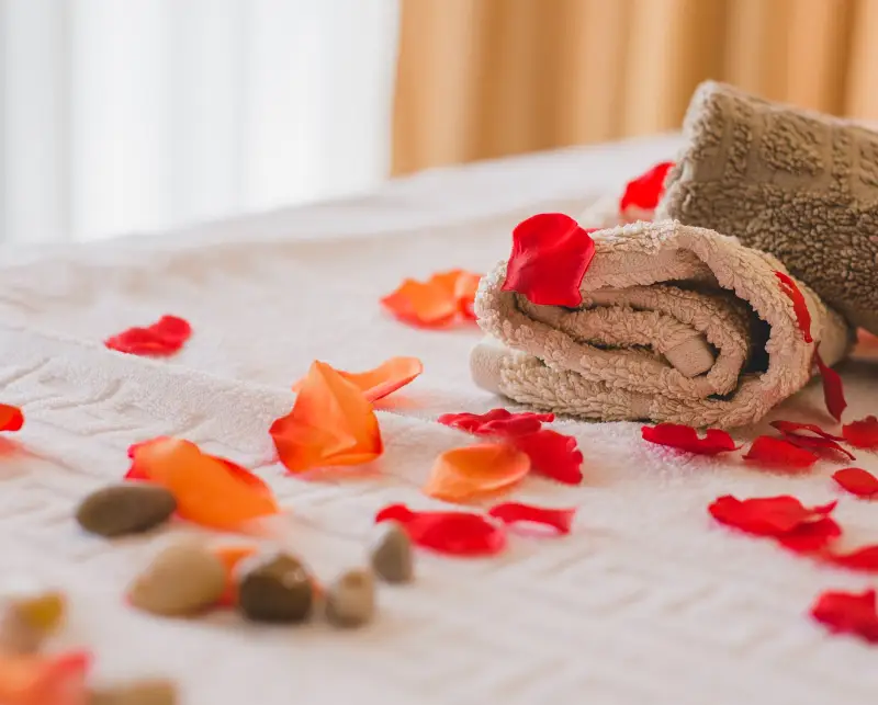 services 034 Soft towels rolled up on a bed scattered with colorful rose petals and smooth stones, creating a serene and inviting spa atmosphere.