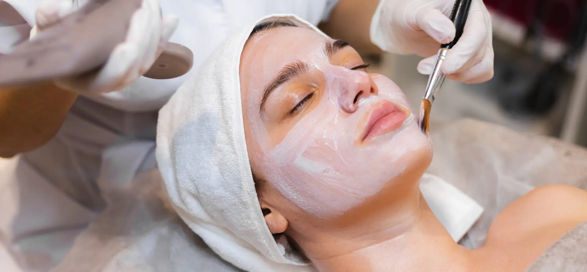 blue webp A person receiving a facial treatment, with hands in gloves applying product using a brush, surrounded by a cozy spa setting.