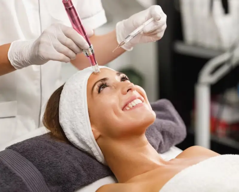 A professional applying a facial treatment using a microneedling device and syringe on a relaxed client lying on a treatment table.